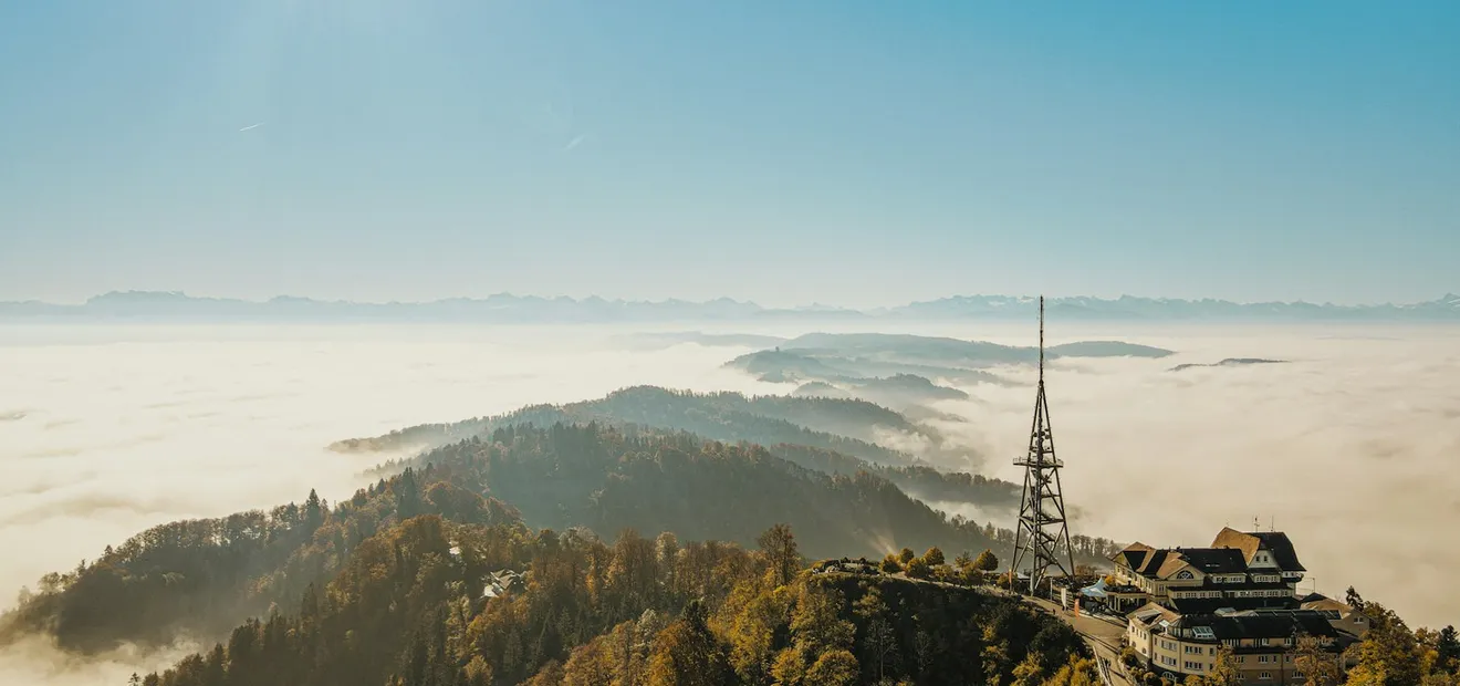 A radio mast and a hilltop building above a sea of fog, with mountains in the distance and the sun high in a clear sky.
