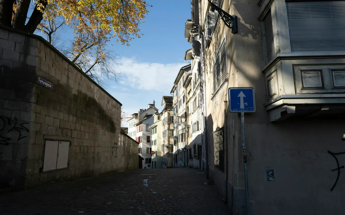 A narrow cobblestone lane between a stone wall and old townhouses, with an autumn tree and blue sky.