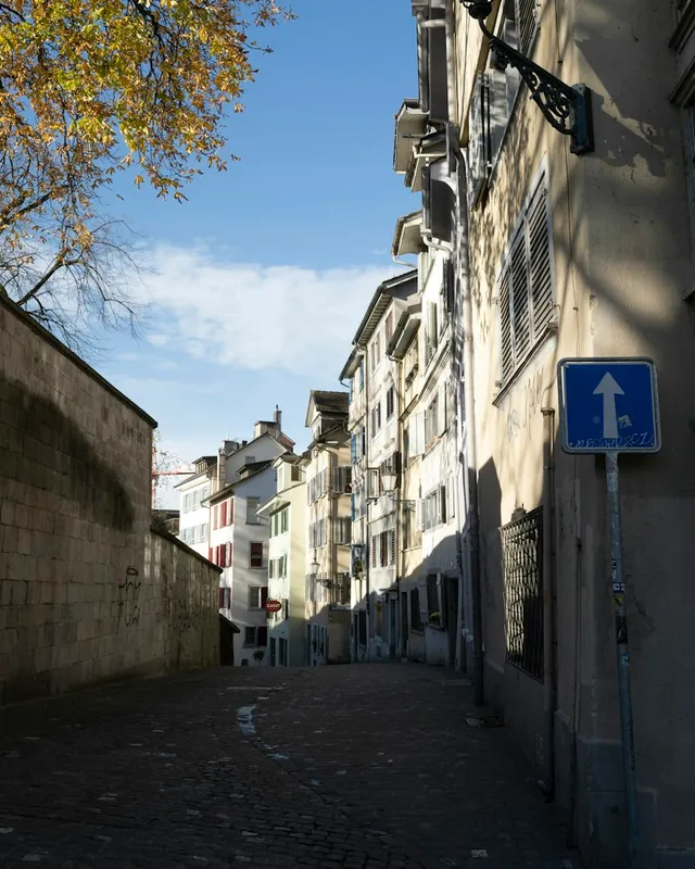 A narrow cobblestone lane between a stone wall and old townhouses, with an autumn tree and blue sky.