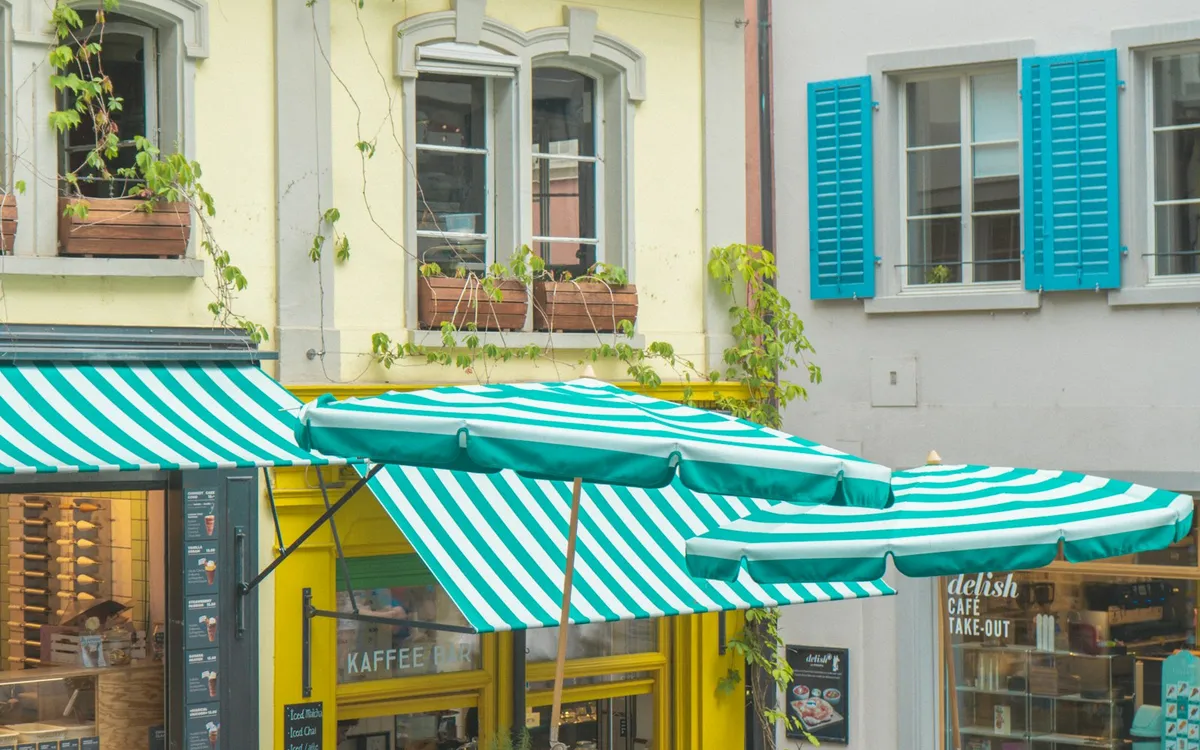 A small café front in a Zurich lane with striped green and white awnings, umbrellas and wooden benches.