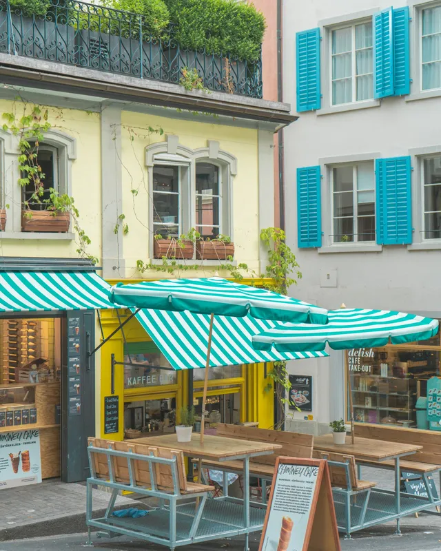 A small café front in a Zurich lane with striped green and white awnings, umbrellas and wooden benches.