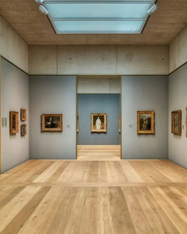 A gallery room with framed paintings on blue-grey walls, a skylight and a wooden floor.